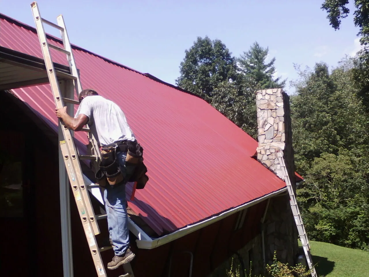 Roofer on a ladder beside a red metal roof and stone chimney — Soffit & Fascia Repair by Roofing Brothers in Sabillasville